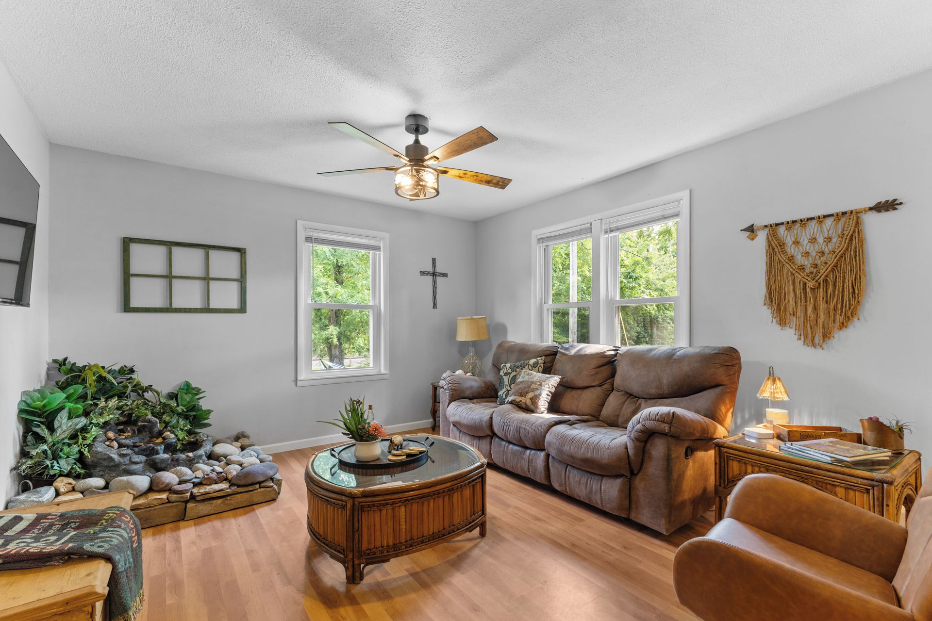 A living room with a brown leather sofa, a glass coffee table, and a ceiling fan. There are two windows with white frames, a cross on the wall, and a macrame wall hanging.