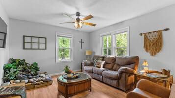A living room with a brown leather sofa, a glass coffee table, and a ceiling fan. There are two windows with white frames, a cross on the wall, and a macrame wall hanging.