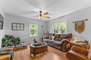 A living room with a brown leather sofa, a glass coffee table, and a ceiling fan. There are two windows with white frames, a cross on the wall, and a macrame wall hanging.