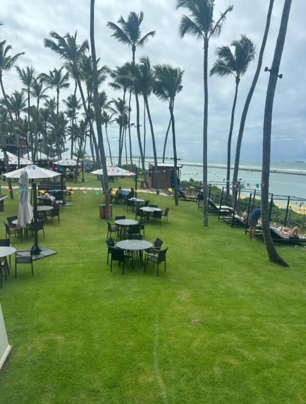 A grassy outdoor area with tables and chairs, palm trees, and a view of the ocean. - Apartment in Muro Alto Beach Resort (Ipojuca)