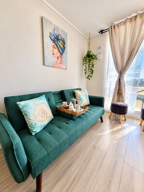 Un sofá color turquesa con una bandeja de objetos, un cuadro en la pared y una ventana con cortinas.