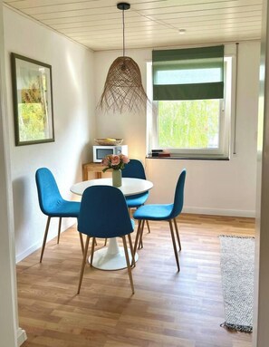 Comedor con una mesa redonda, sillas azules, una lámpara colgante y una ventana con vista a follaje.