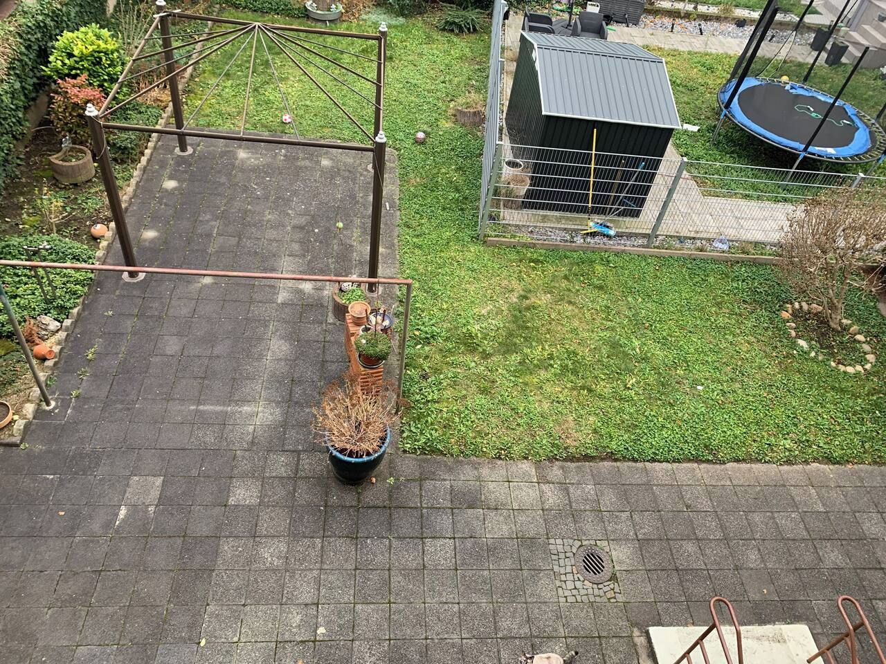 Et beboelsesområde udendørs med en flisebelagt terrasse, en lille have med en potteplante, en trampolin og et skur.
