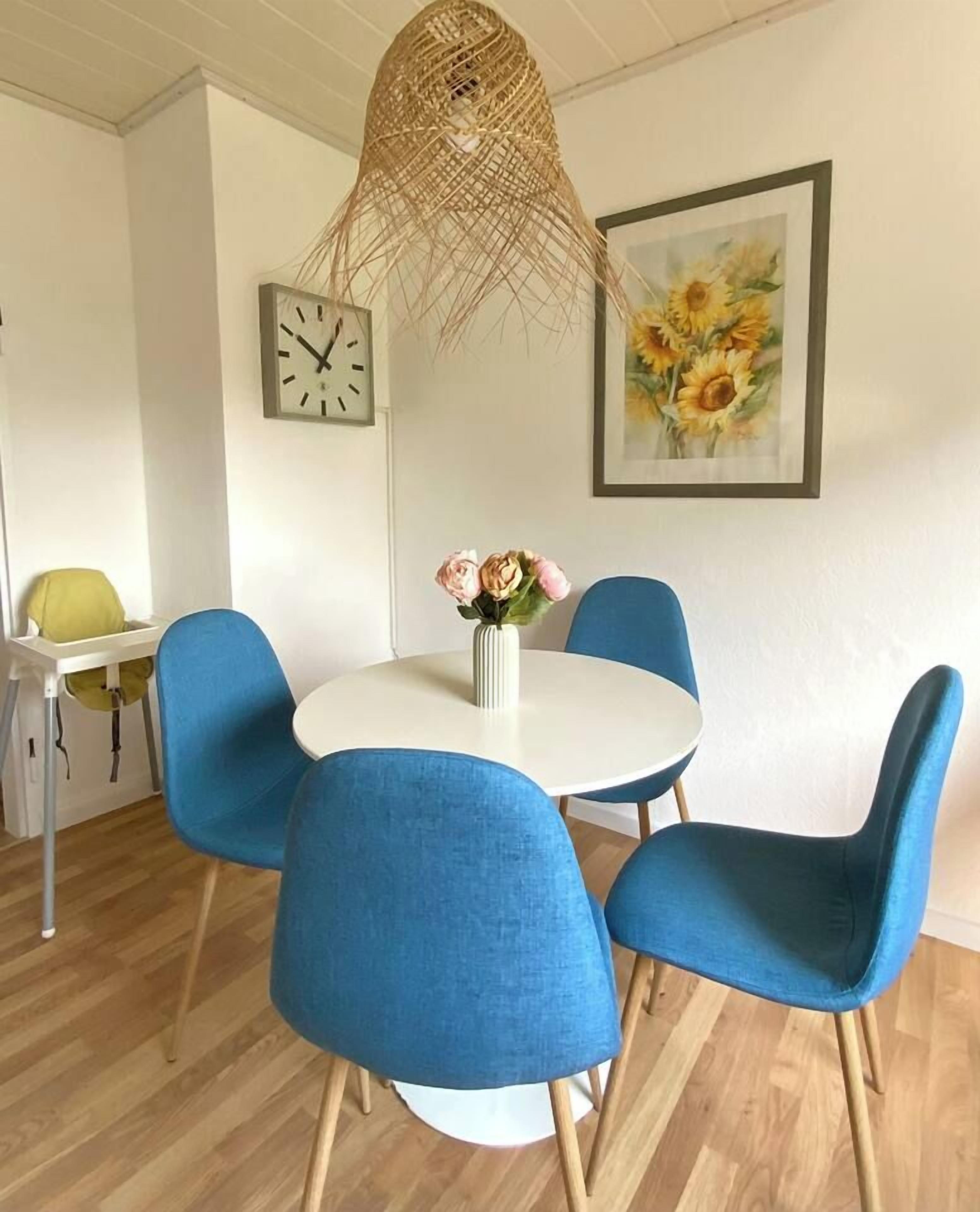 Ein Essbereich mit einem runden Tisch, vier blauen Stühlen, einer Blumenvase, einer Wanduhr und einem Gemälde von einem Sonnenblumenfeld.