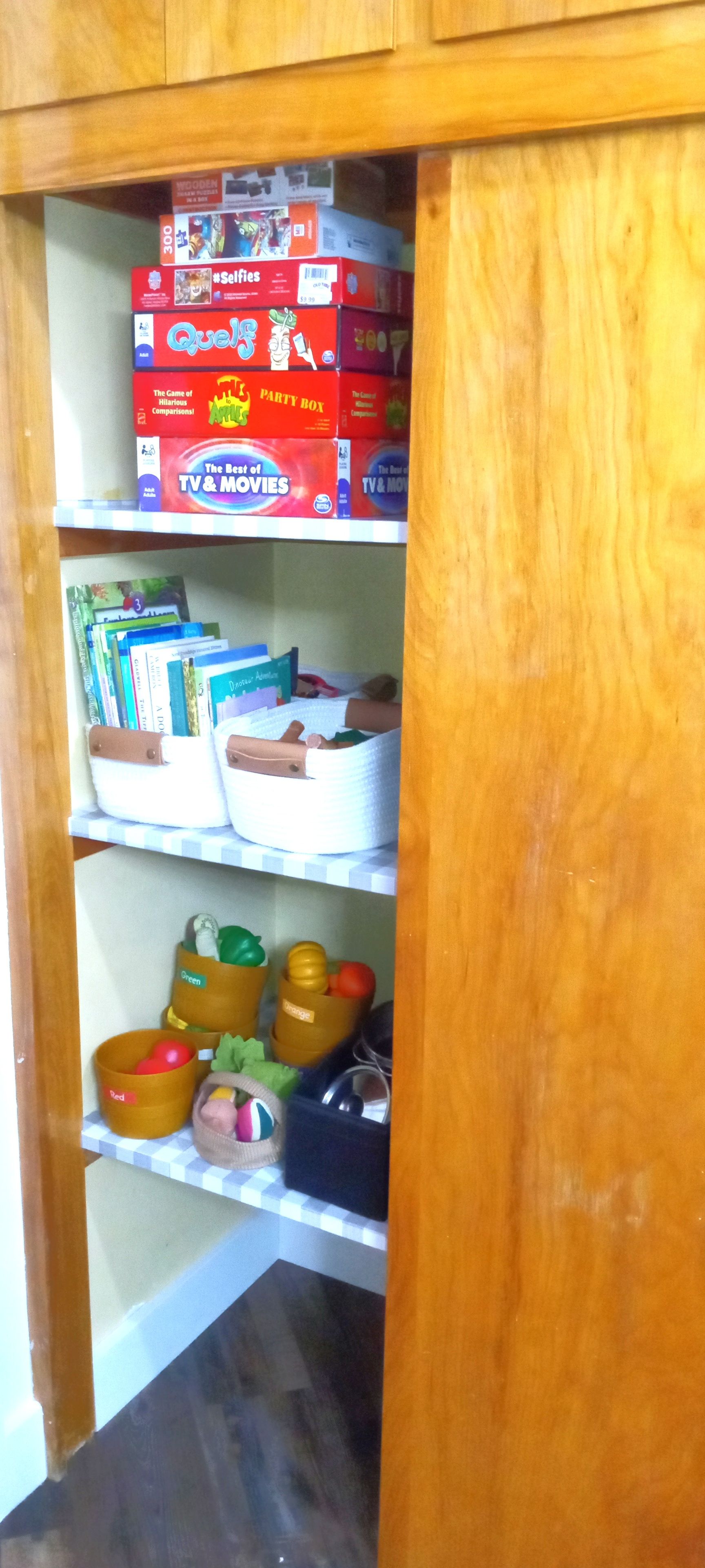 A wooden cabinet with shelves containing various items including books, a board game, and storage bins.