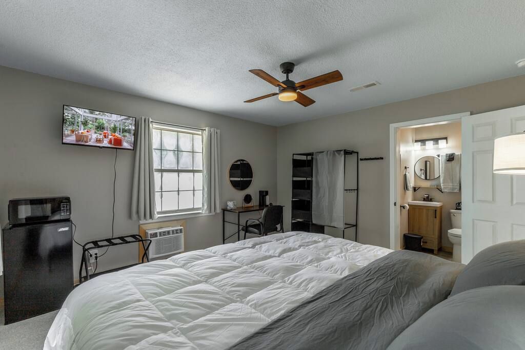 A bedroom with a bed, a ceiling fan, a television, a desk, a chair, a window, and a bathroom.