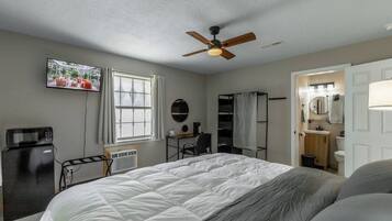 A bedroom with a bed, a ceiling fan, a television, a desk, a chair, a window, and a bathroom.