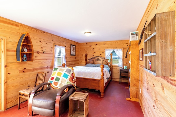A cozy wooden room with a bed, a chair, a small table, and a window.
