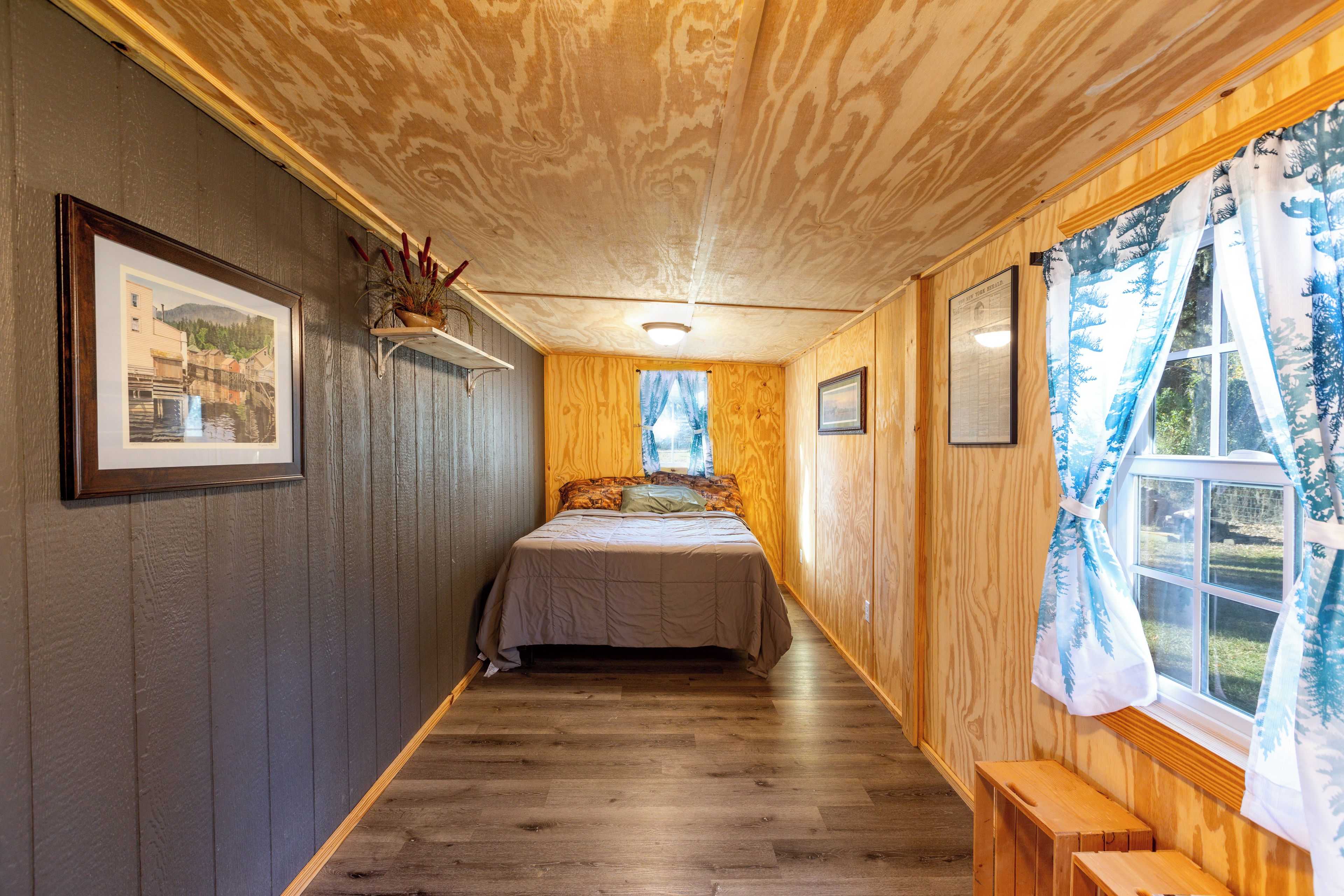 Un estrecho pasillo que lleva a una cama, con suelo y paredes de madera, y una ventana con cortinas.