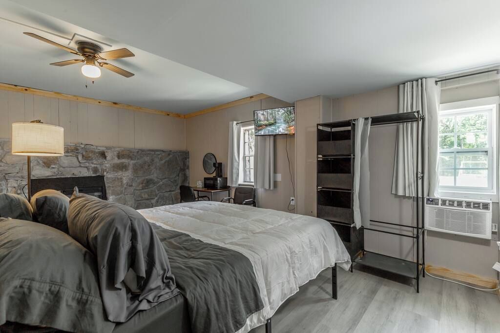 A bedroom with a bed, a ceiling fan, a stone wall, a TV, a desk, and a window with curtains.