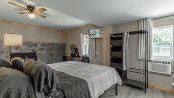 A bedroom with a bed, a ceiling fan, a stone wall, a TV, a desk, and a window with curtains.