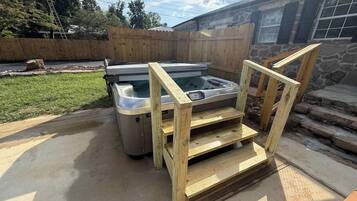 A hot tub with a wooden frame and steps, located in a backyard with a stone building and a wooden fence in the background.