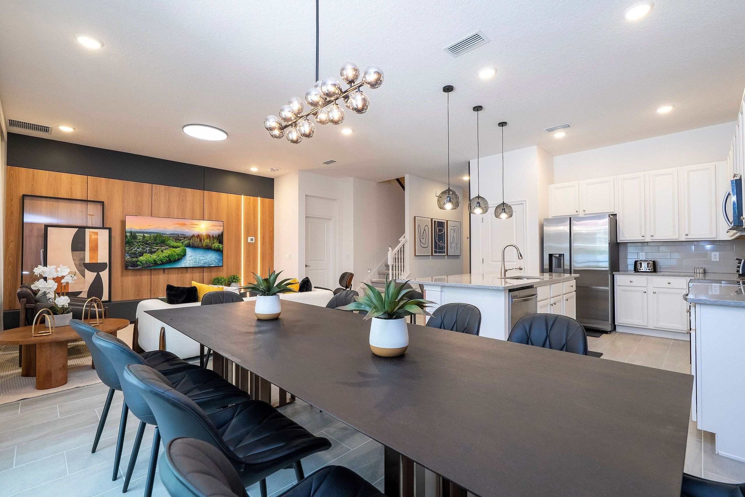 A modern kitchen with stainless steel appliances, a dining area with a long table and chairs, and a living space with a TV and wooden paneling.