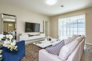 Ein modernes Wohnzimmer mit Fernseher, Sofa, Couchtisch und einem großen Fenster mit Jalousien.