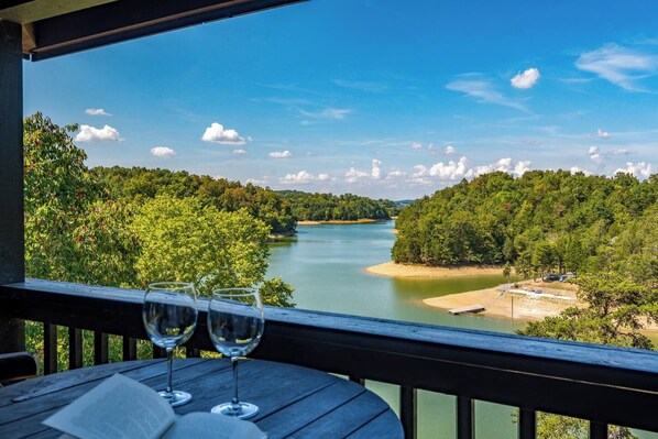 A wooden deck with a table and two wine glasses, overlooking a river and forested area. - Lakefront at Douglas Lake - Private Dock - Hot Tub (Sevierville)