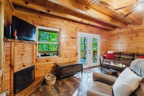 L’intérieur d’un chalet en bois est aménagé avec un téléviseur, une cheminée, un canapé et offre une vue sur la verdure environnante.