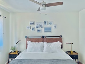 Une chambre à coucher avec un lit, un ventilateur de plafond, des tables de chevet avec des lampes et des œuvres d’art encadrées accrochées au mur.
