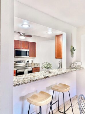 A kitchen with a granite countertop, stainless steel appliances, wooden cabinets, and two bar stools. - <2 min to Beach! Kihei Condo w/ Ocean View and AC (Kihei)