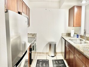 A compact kitchen with a stainless steel refrigerator, granite countertop, wooden cabinets, and a stainless steel trash can. - <2 min to Beach! Kihei Condo w/ Ocean View and AC (Kihei)
