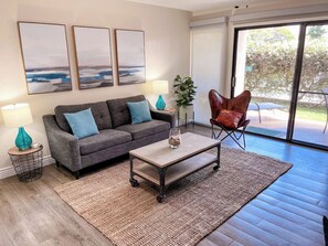 Ein modernes Wohnzimmer mit einem grauen Sofa, einem Couchtisch und einem roten Sessel. An der Wand hängen drei gerahmte Kunstwerke, eine Pflanze und ein großer Fensterblick.