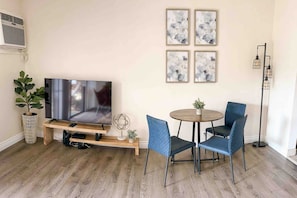 A living room with a flat-screen TV on a wooden stand, a round table with chairs, and a floor lamp.