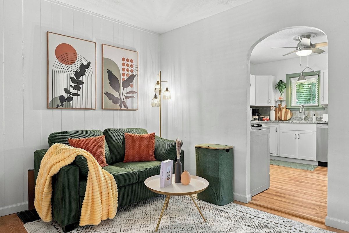 A living room with a green sofa, a round coffee table, and a small side table. There is a kitchen visible through an archway.