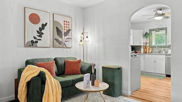 A living room with a green sofa, a round coffee table, and a small side table. There is a kitchen visible through an archway.