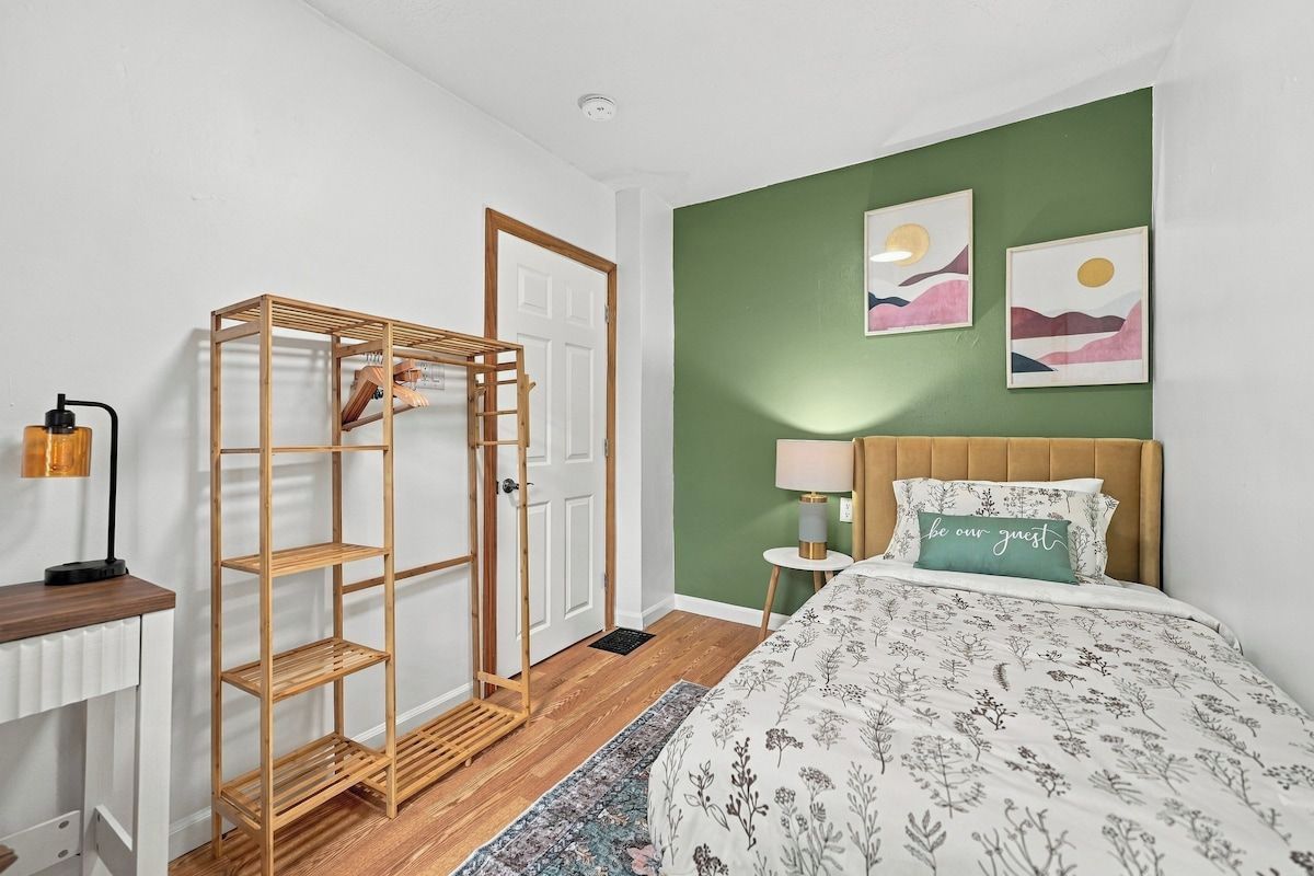 A bedroom with a bed, a wooden wardrobe, a bedside table with a lamp, and a green wall with artwork.