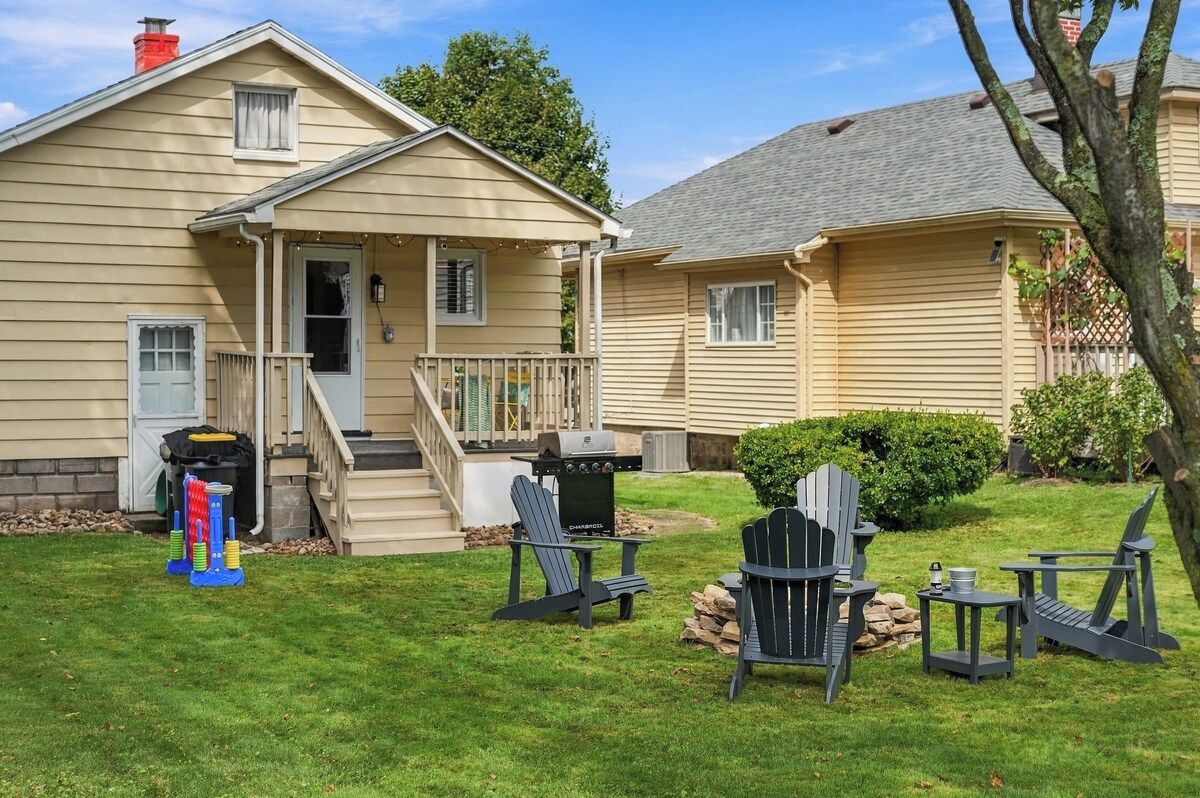 Una casa de dos pisos con un porche delantero, una parrilla y mobiliario exterior con sillas tipo Adirondack.