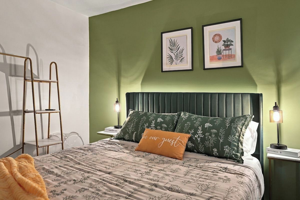 A neatly made bed with a floral-patterned bedspread and a single orange pillow with text.
