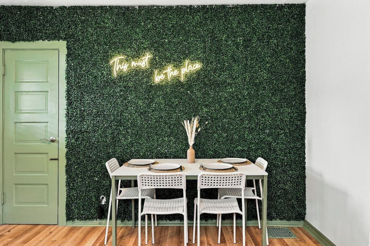 A dining area with a table set for four, a green door, and a wall with a green leafy backdrop and a neon sign.