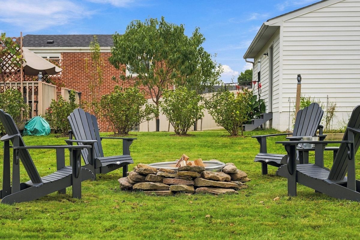 Een achtertuin met twee Adirondack stoelen, een vuurkorf van steen en een huis met bakstenen.