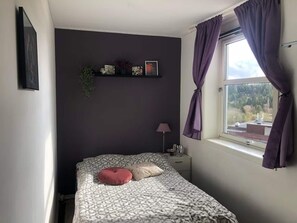A bedroom with a bed, a nightstand, a lamp, a window with curtains, and wall-mounted pictures. - Apartment in Romolslia (Trondheim)