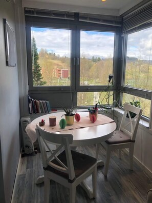 A room with a round table, four chairs, a radiator, and large windows with a view of trees and buildings. - Apartment in Romolslia (Trondheim)