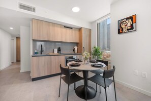 A modern kitchen with wooden cabinets, a dining area with a round table and black chairs, and a view of a cityscape through a large window. - 501 First Corner Balcony City Views (Miami)