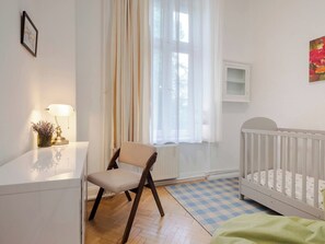 Ein Zimmer mit einem Babybett, einem Sessel, einem Schreibtisch, einer Lampe und einem fenster mit Vorhängen.