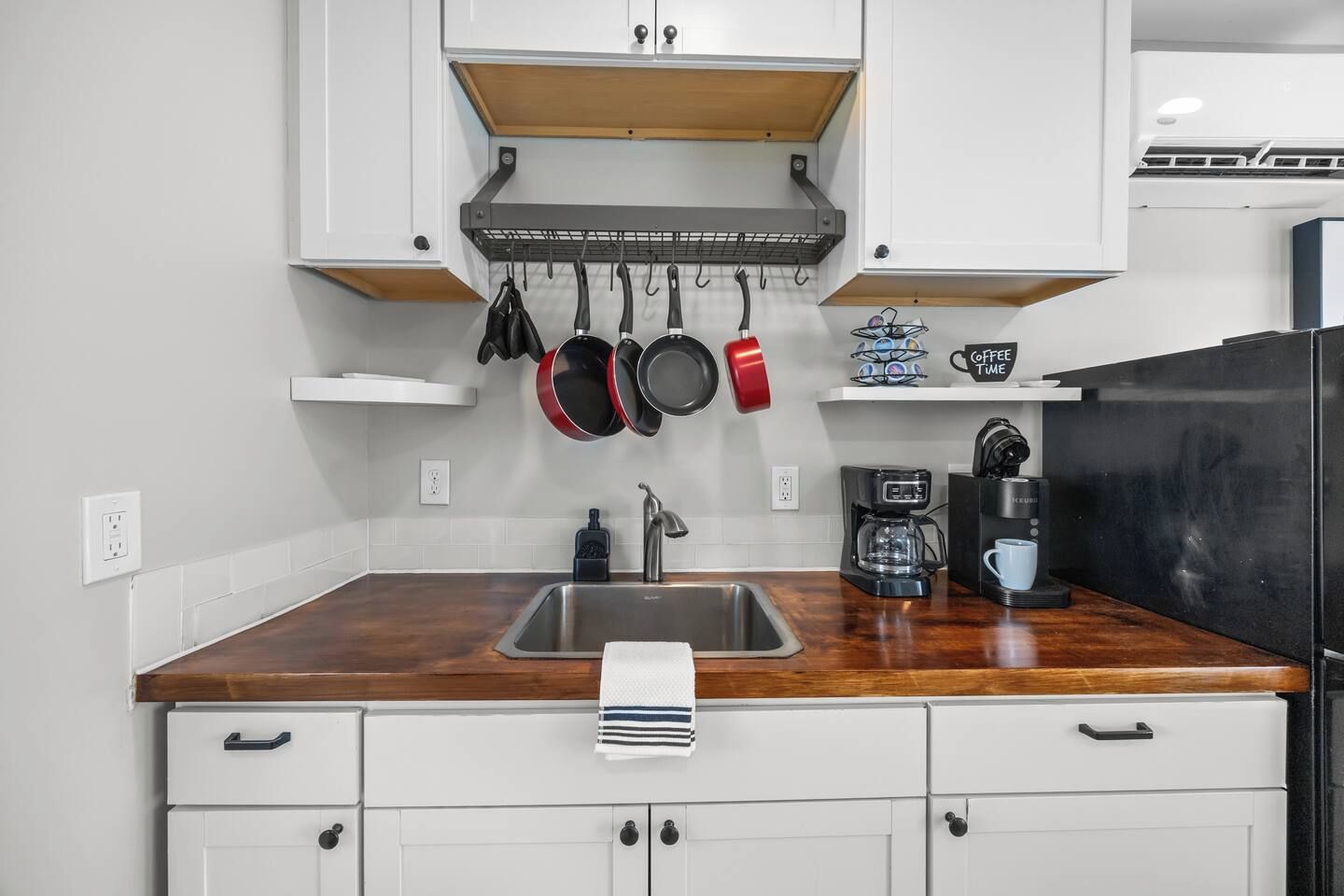 A kitchen with a wooden countertop, a stainless steel sink, hanging pots, a coffee maker, and white cabinets.