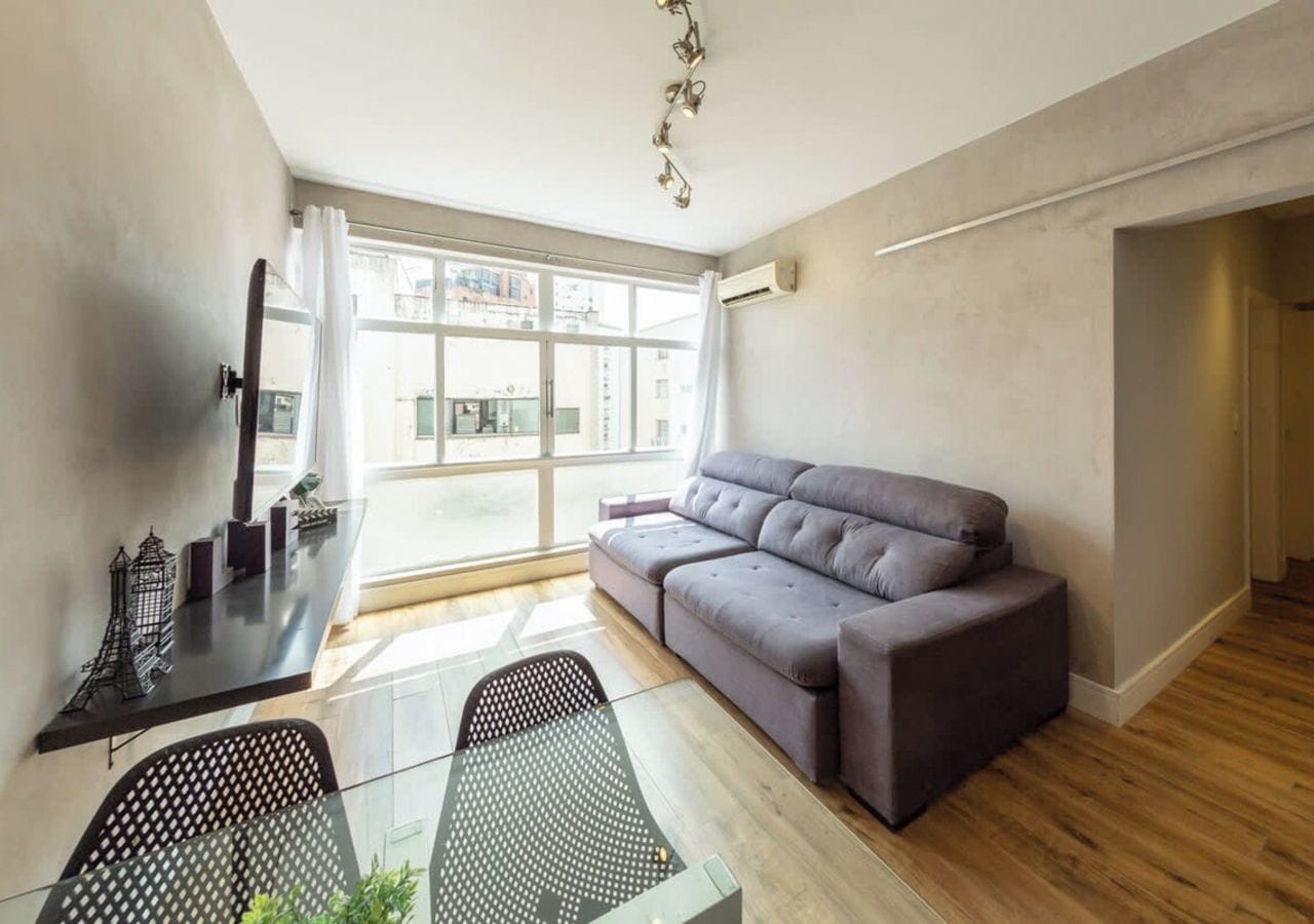 A modern living room with a grey sofa, a glass dining table, and a city view through large windows.