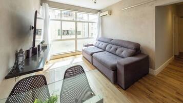 Ett modernt vardagsrum med en grå soffa, ett glasbord och utsikt över staden genom stora fönster.