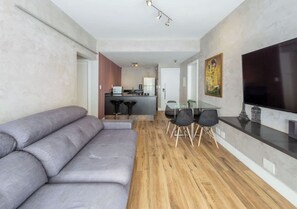 Een moderne woonkamer met een grijze hoekbank, een eethoek met een glazen tafel en stoelen, en een keuken op de achtergrond.