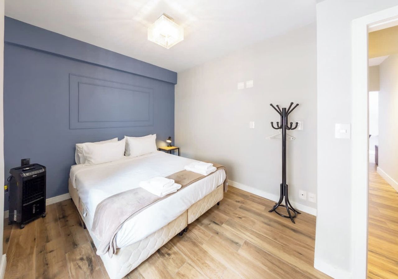 A bedroom with a bed, bedside table, lamp, coat rack, and a suitcase.