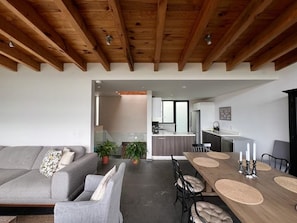 Ein modernes Wohnzimmer mit einer Couch, einem Esstisch und Stühlen. Im Hintergrund ist eine Küchenzeile mit einem Kühlschrank und Schränken zu sehen.