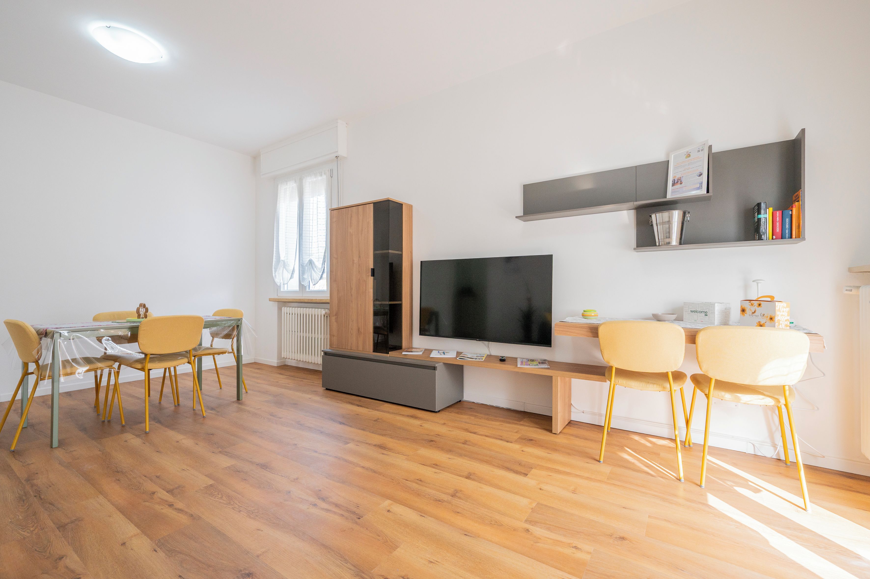 A modern living room with a flat-screen TV, wooden flooring, and a dining area with yellow chairs.