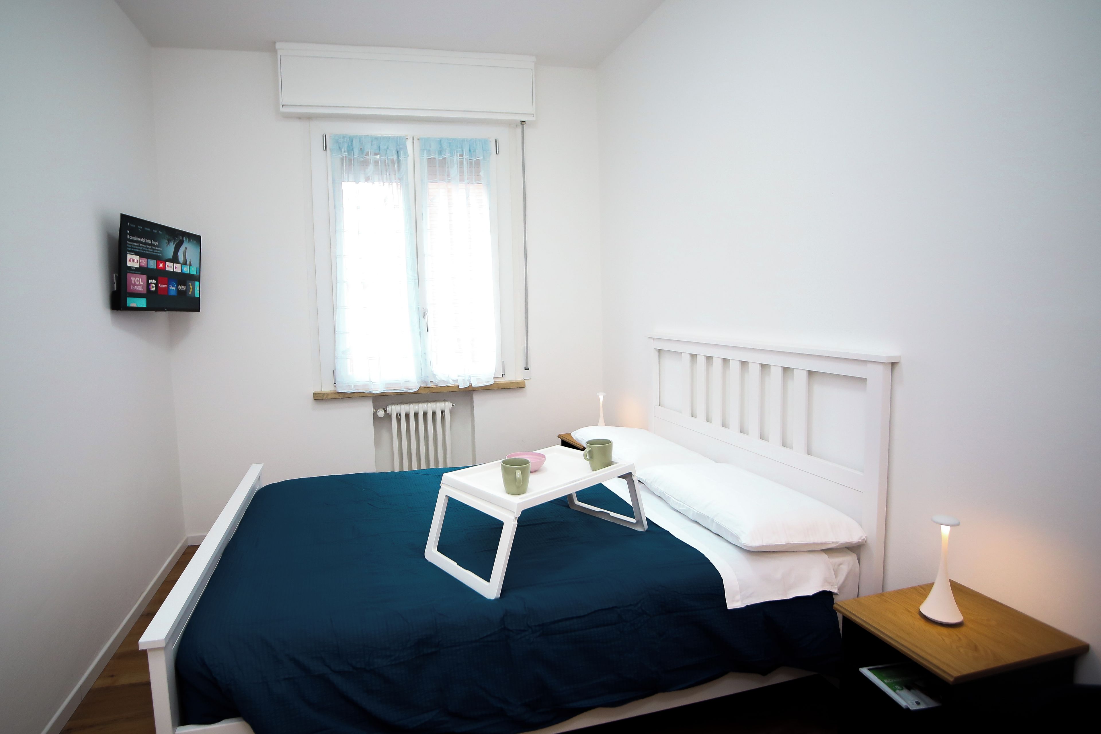 A small, neatly arranged bedroom with a single bed, a bedside table with a lamp, and a wall-mounted TV.
