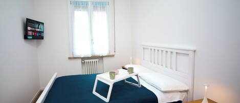 A small, neatly arranged bedroom with a single bed, a bedside table with a lamp, and a wall-mounted TV.