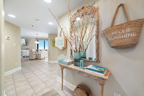 A hallway with a console table displaying books and decorative items, a mirror with a 'Hello Sunshine' tote bag, and a dining area in the background. - "Broadmoor #705" --- New! Beachfront 3BR, Stunning Gulf Views, Pool, Hot Tub (Orange Beach)