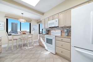 A modern kitchen with a built-in oven, microwave, and refrigerator. There is a dining area with a table and chairs, and a view of a cityscape through large windows. - "Broadmoor #705" --- New! Beachfront 3BR, Stunning Gulf Views, Pool, Hot Tub (Orange Beach)