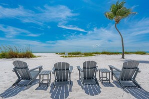 A beach with four Adirondack chairs and a table, a palm tree, and a clear blue sky. - The Turtle Nest (Fort Myers Beach)