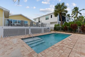 A swimming pool with a white deck, surrounded by a fence and palm trees. - Oasis on Ohio Ave (Fort Myers Beach)