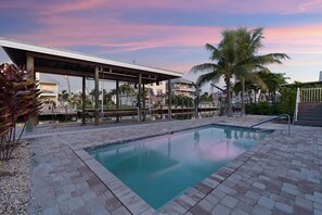 A swimming pool with a brick border, surrounded by palm trees and a covered area with a view of a residential building. - Oasis on Ohio Ave (Fort Myers Beach)